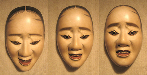 Three human-face masks handing on the wall. Each has a different facial expression showing a different emotion. Three pictures of the same mask. A different facial expression shows on the mask depending on which angle you view the mask.