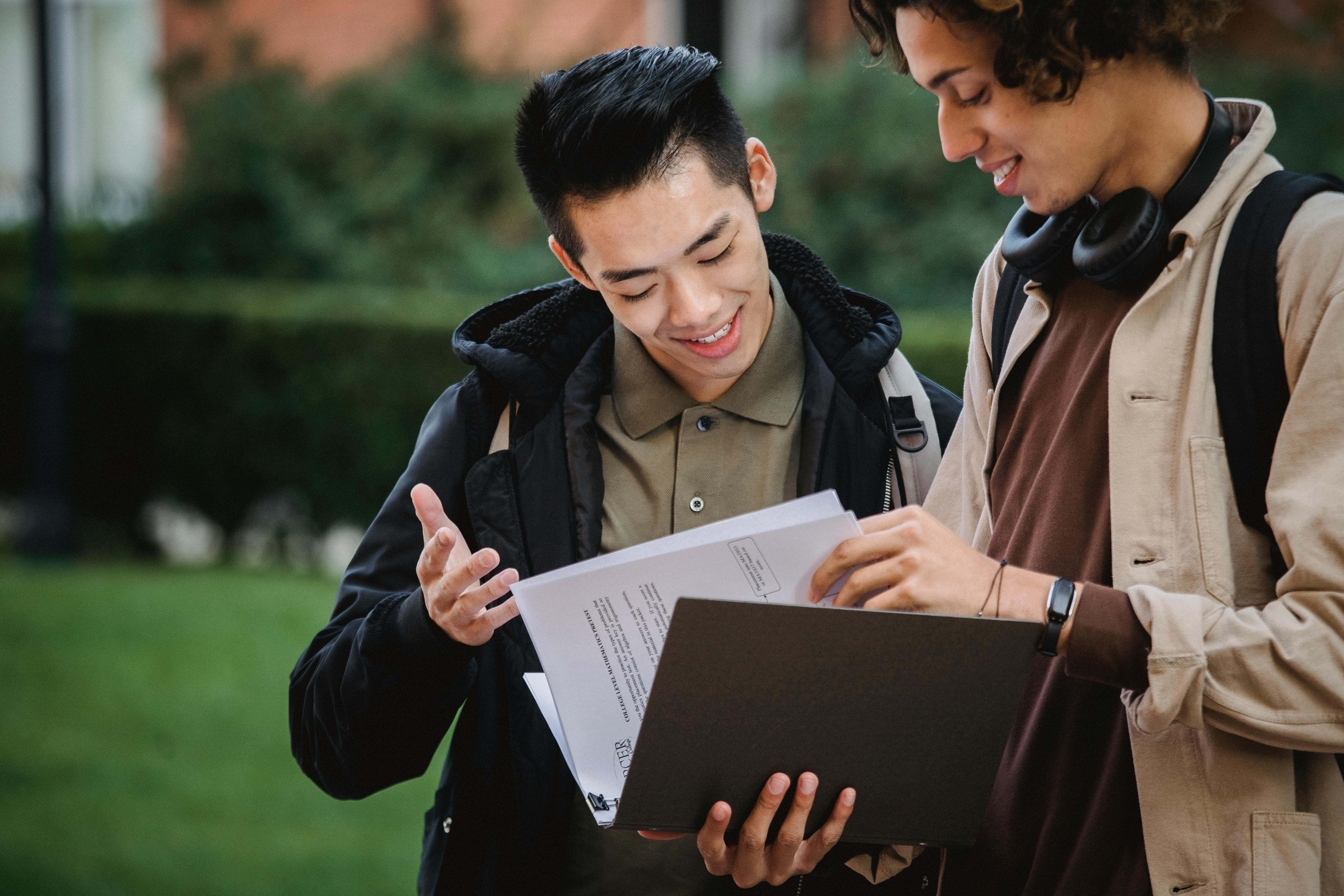 A university student and his teaching assistant smiling and chatting on the school lawn. The student is showing the TA his assignment paper, which is held in his binder.