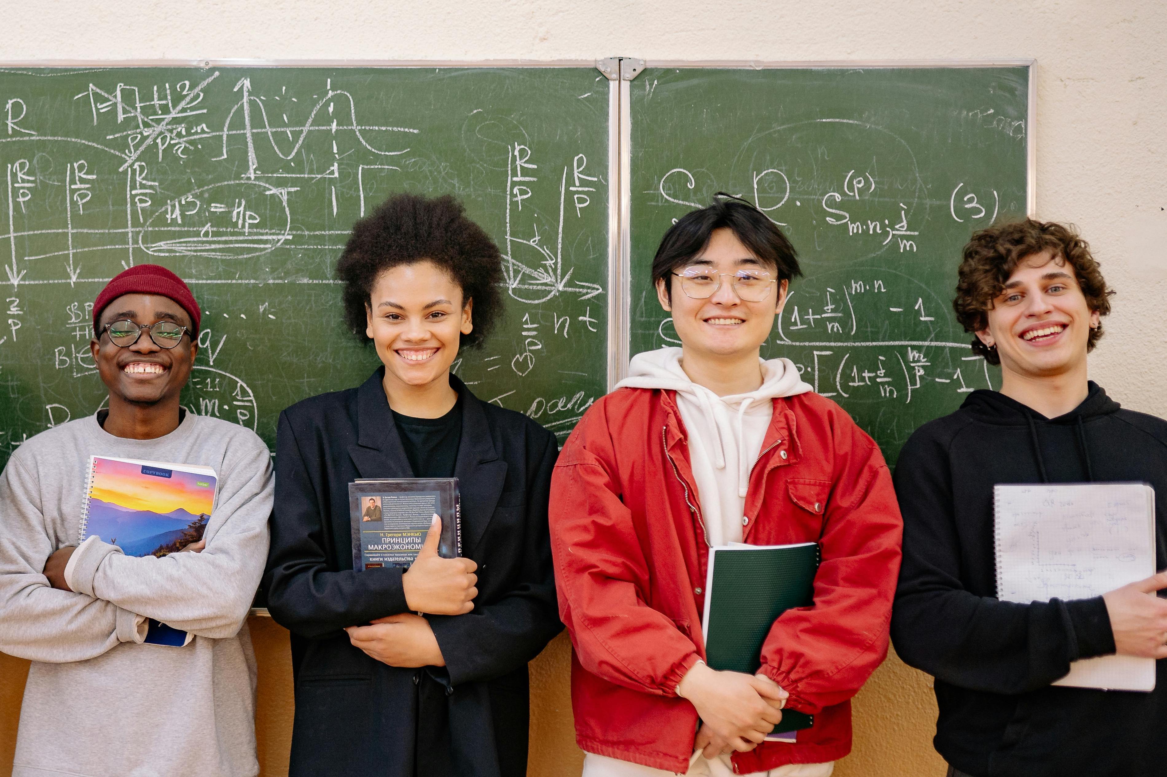 A group of diverse young adults standing in front of a blackboard with mathematical equations on it. They are holding notebooks in their hands.