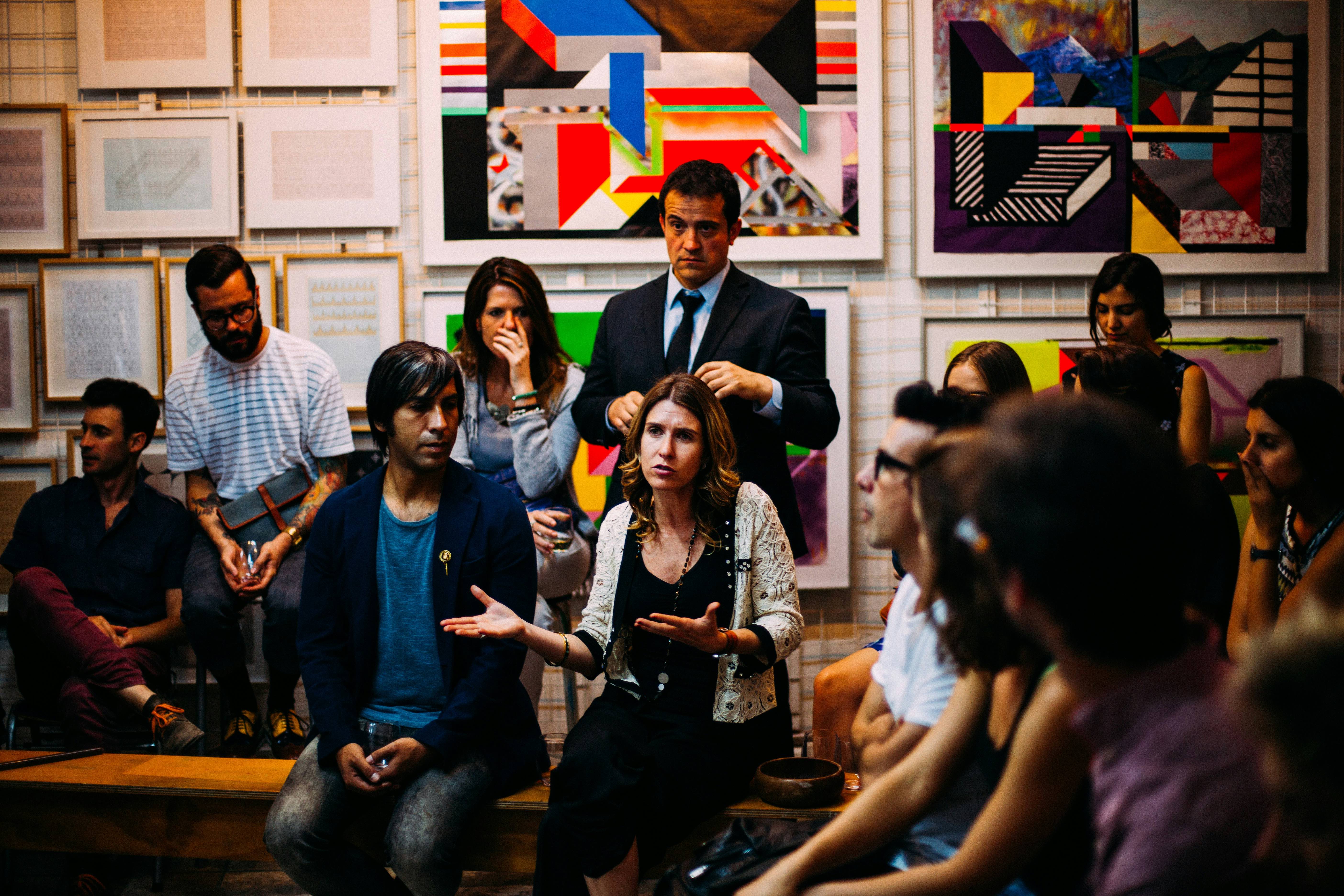 A class discussion. A woman is seated in a chair and talking. Others in the room are sitting around her in a circle, or standing behind.