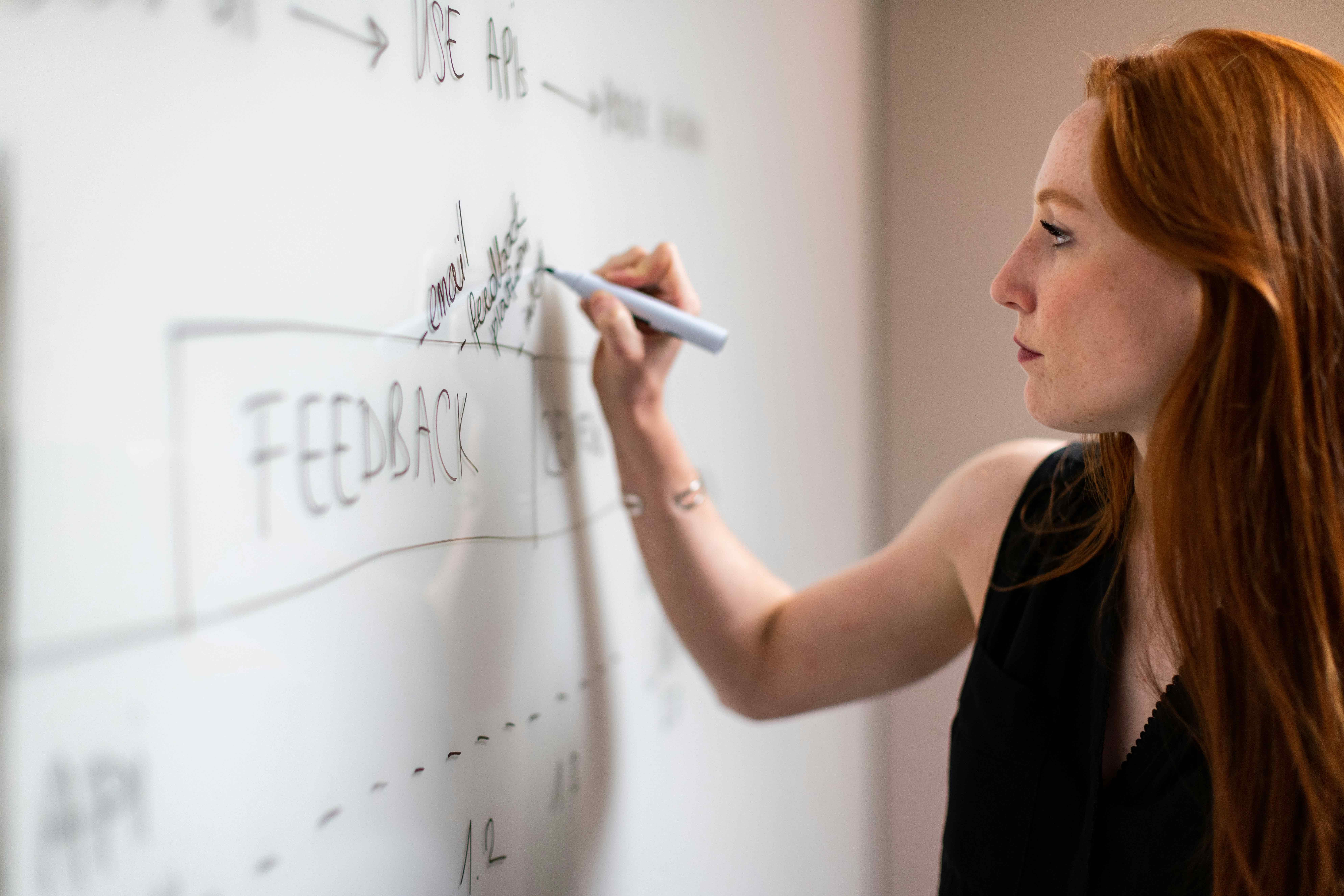 A female teaching assistant writing on the board. There are words such as 'use APIs', 'Feedback' and 'Email' on the board