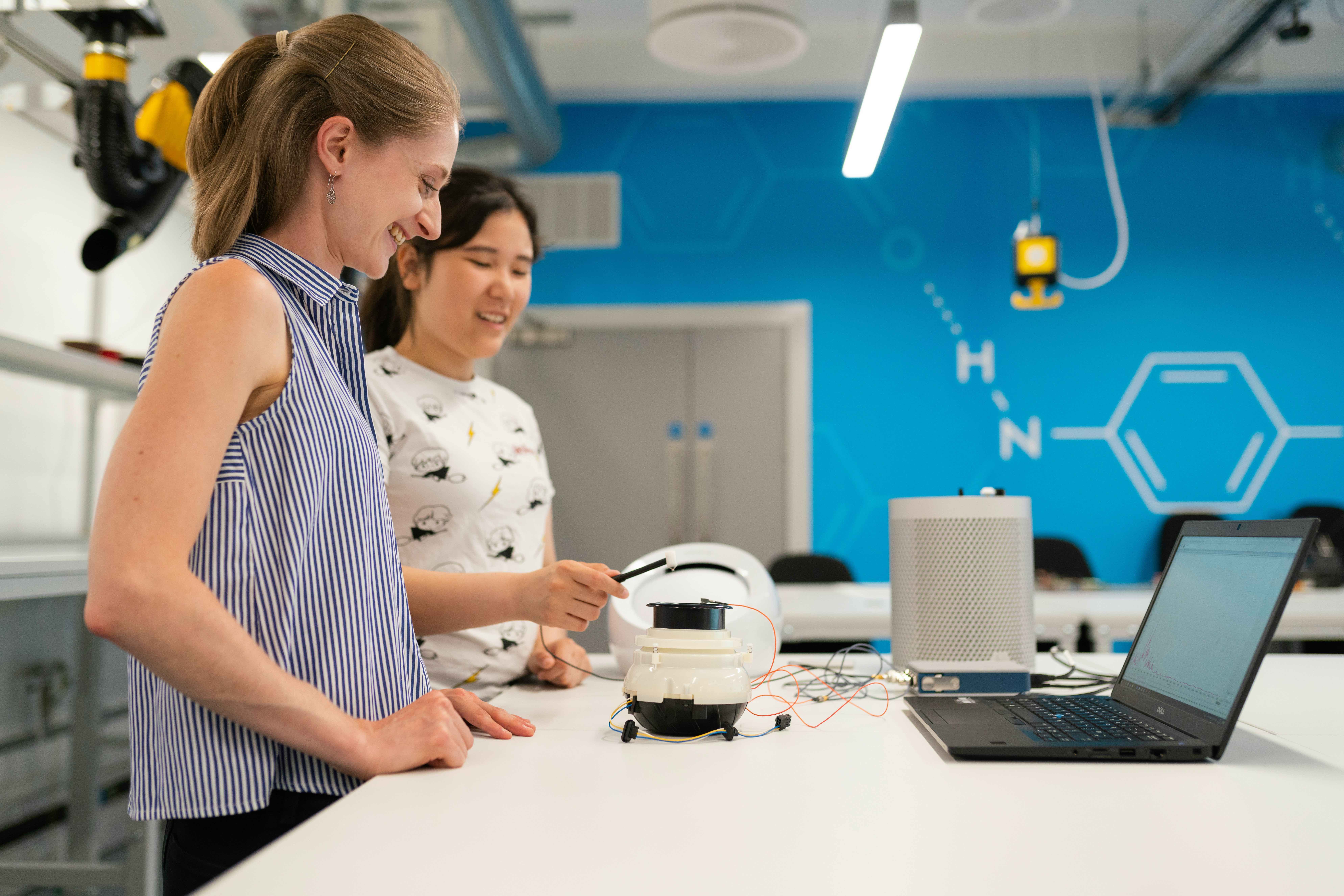 A photo of a teaching assistant with a student. They are working on a robotics project in a laboratory setting. The TA is helping the student with an electronic device that is connected to their laptop.