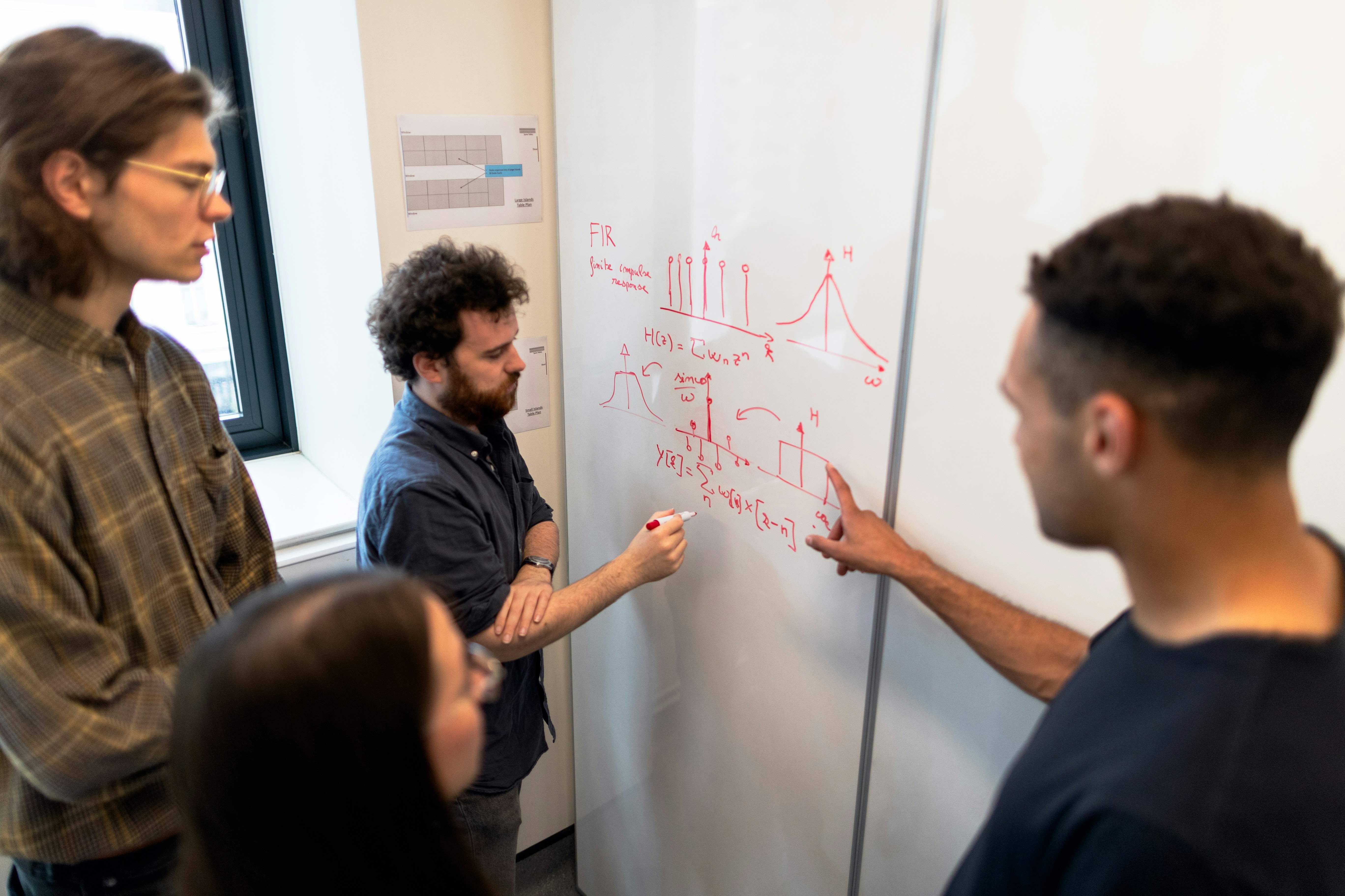 A group of students gathered around a whiteboard, and the instructor writing mathematical equations with a red marker on it.