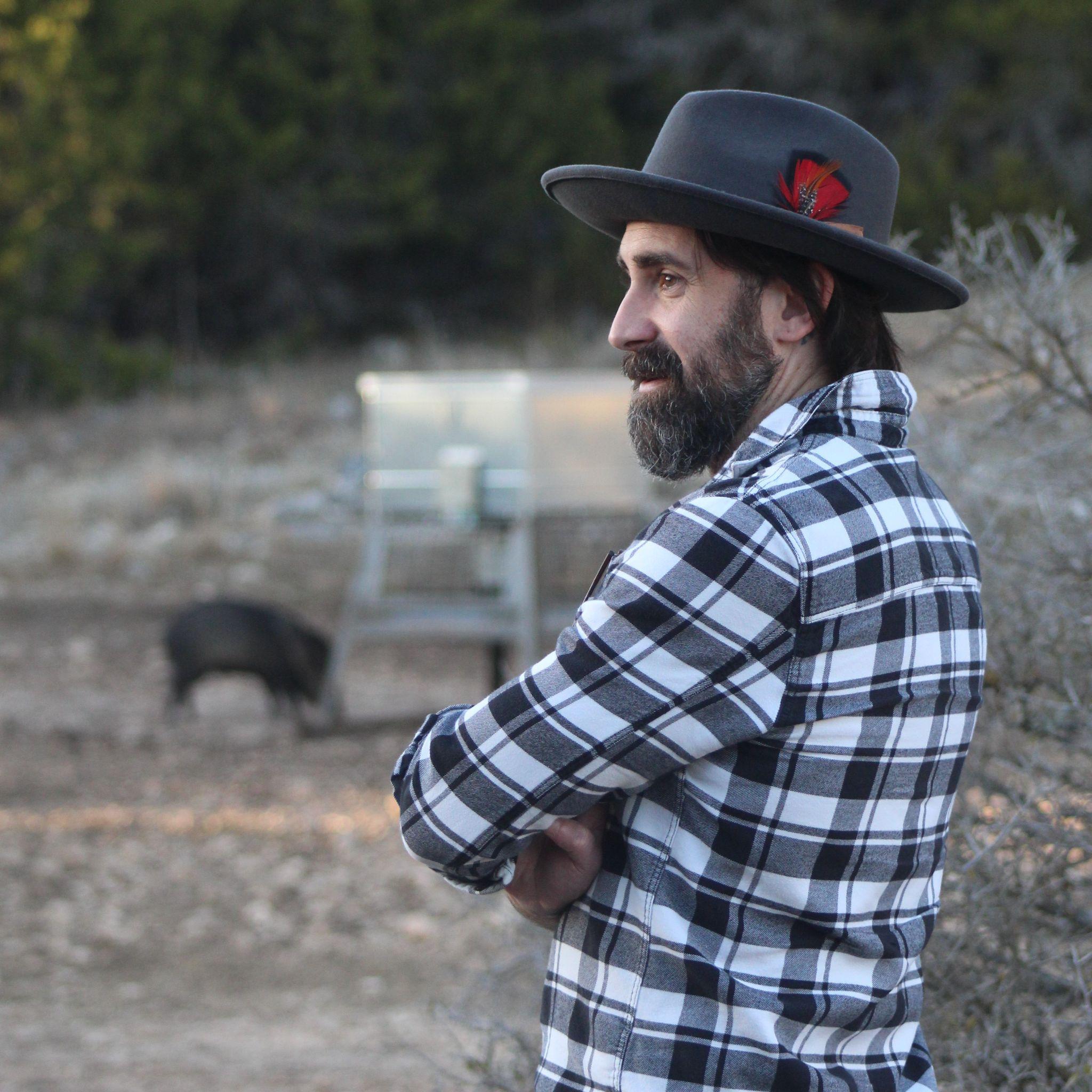 A bearded man wearing a fedora hat looks off in the distance. 