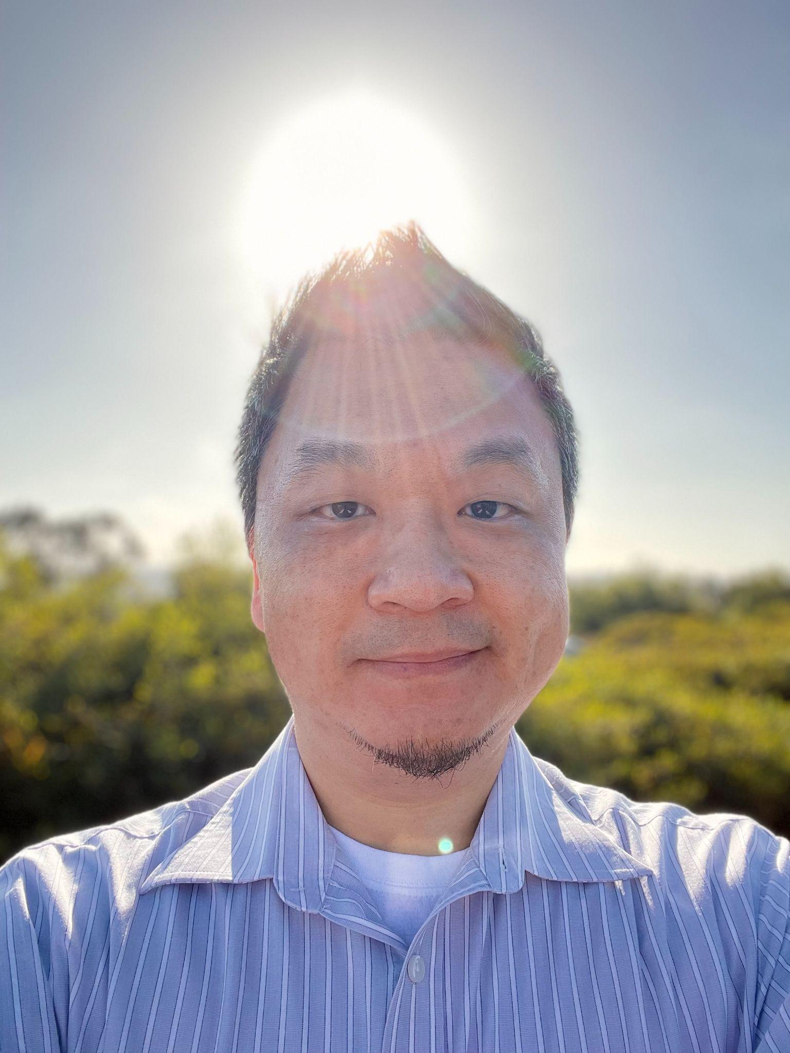 A man with short, black hair stands outdoors with a setting sun behind him.