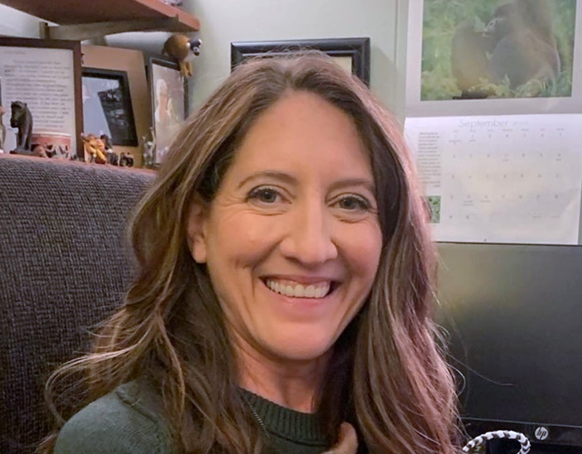 Headshot of smiling person with longer brown hair. Picture frames are in the background.