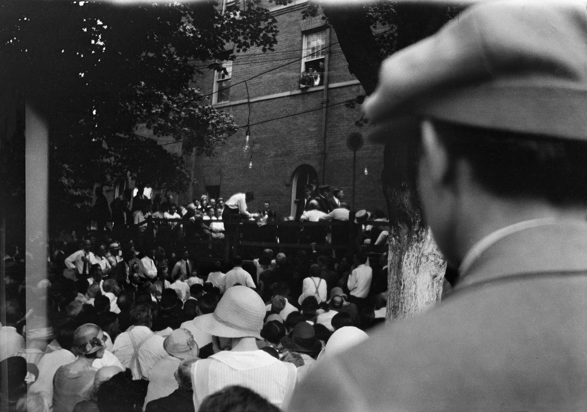 A crowd of people face a fenced-off area outside a brick building.