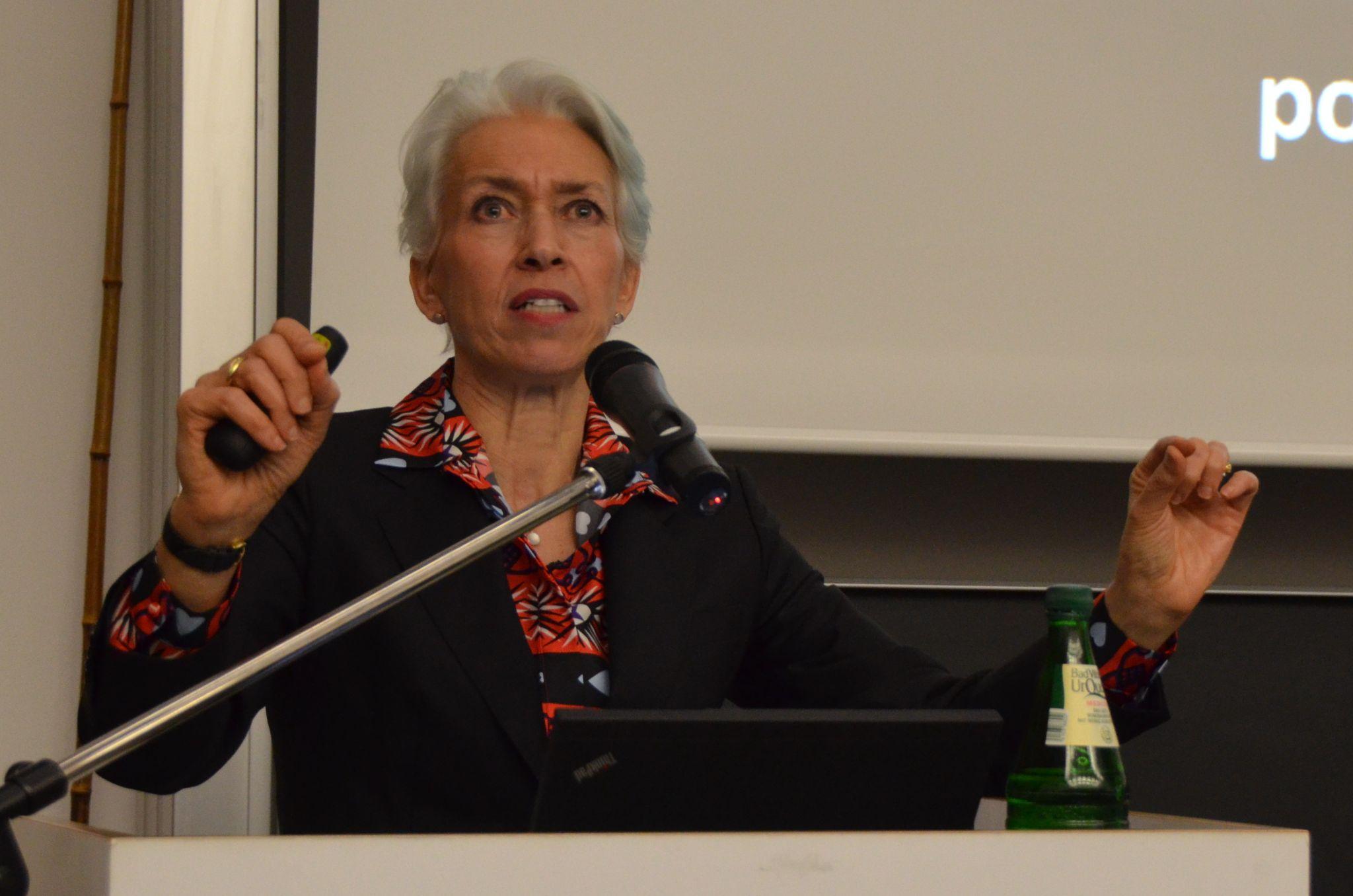 Person standing at podium in front of a screen with arms partly raised.